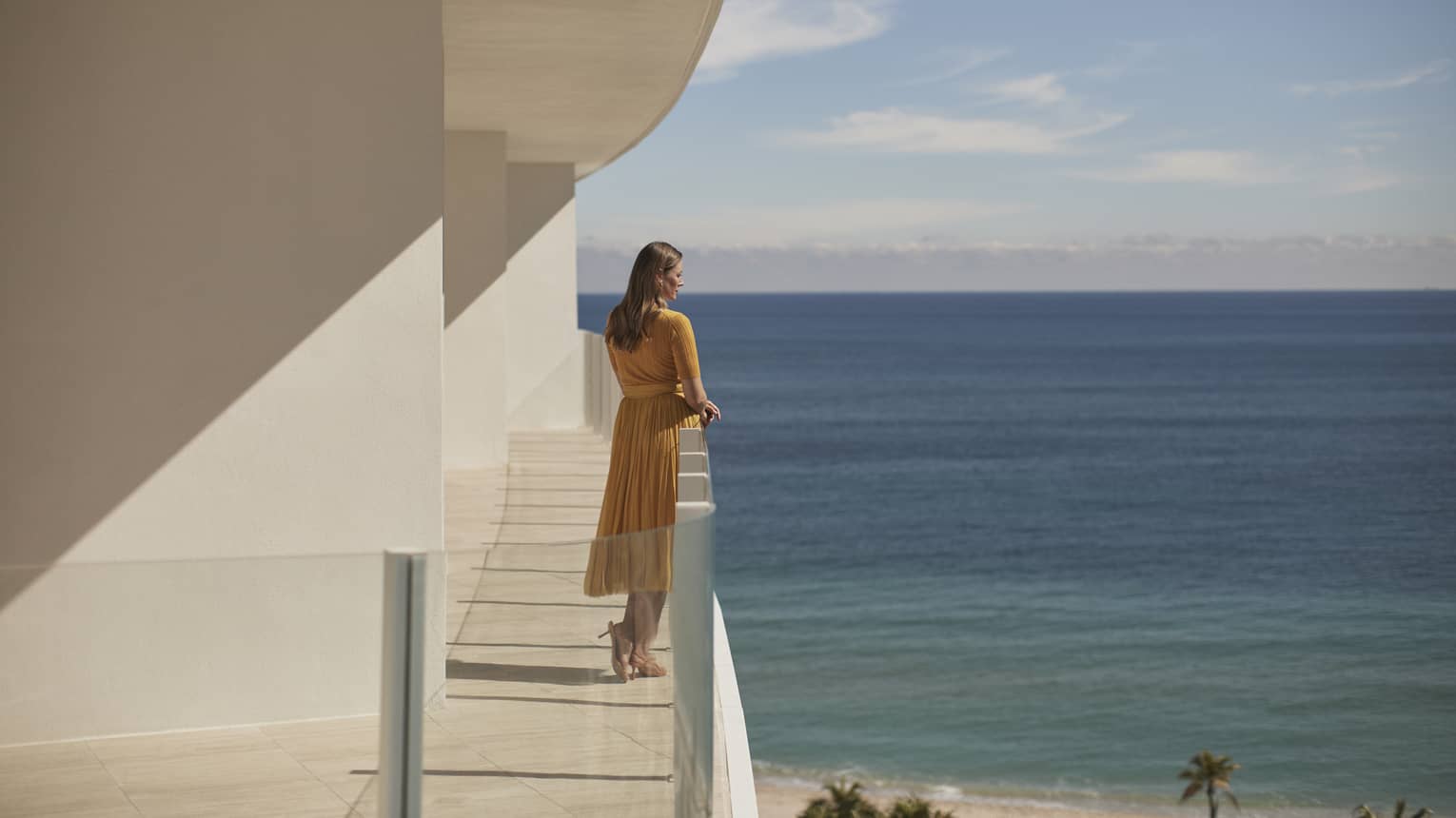A woman standing on a balcony  of a white stone building with the ocean in the background.