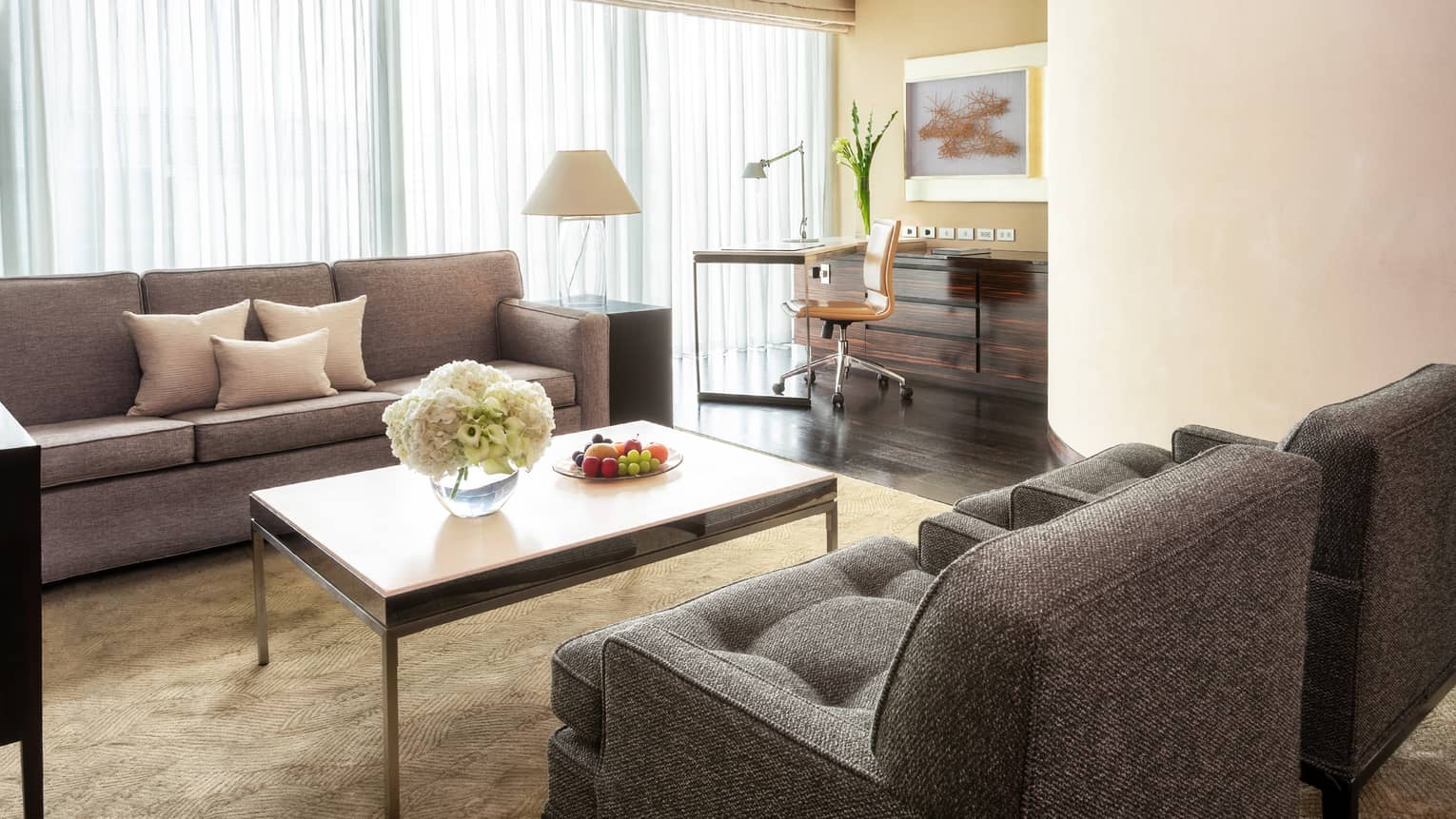 Living area with grey sofas, coffee table with flowers and fruit, work desk with a chair and floor-to-ceiling windows with curtains