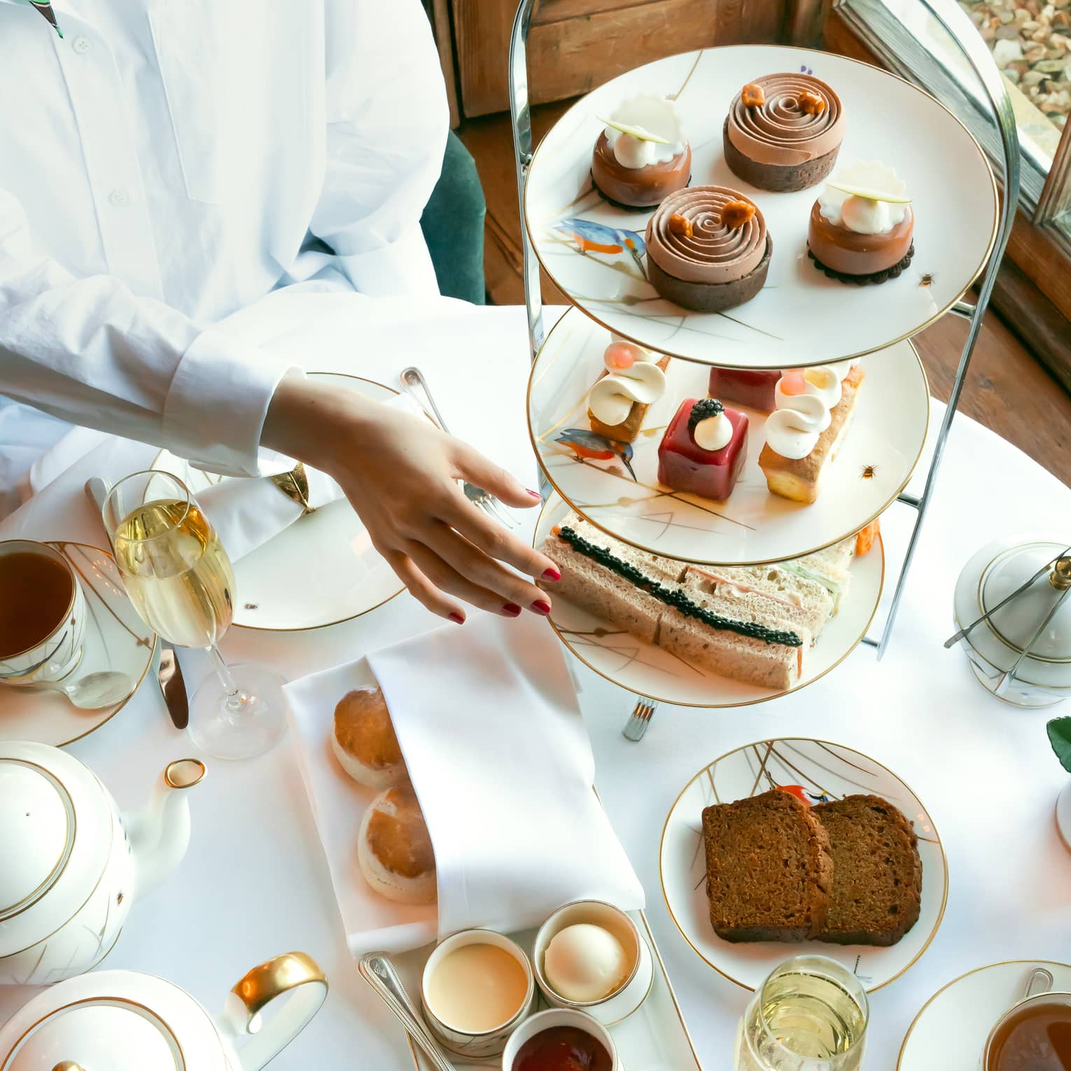 Person in white shirt reaches to classic afternoon tea stand filled with patisserie and mini sandwiches