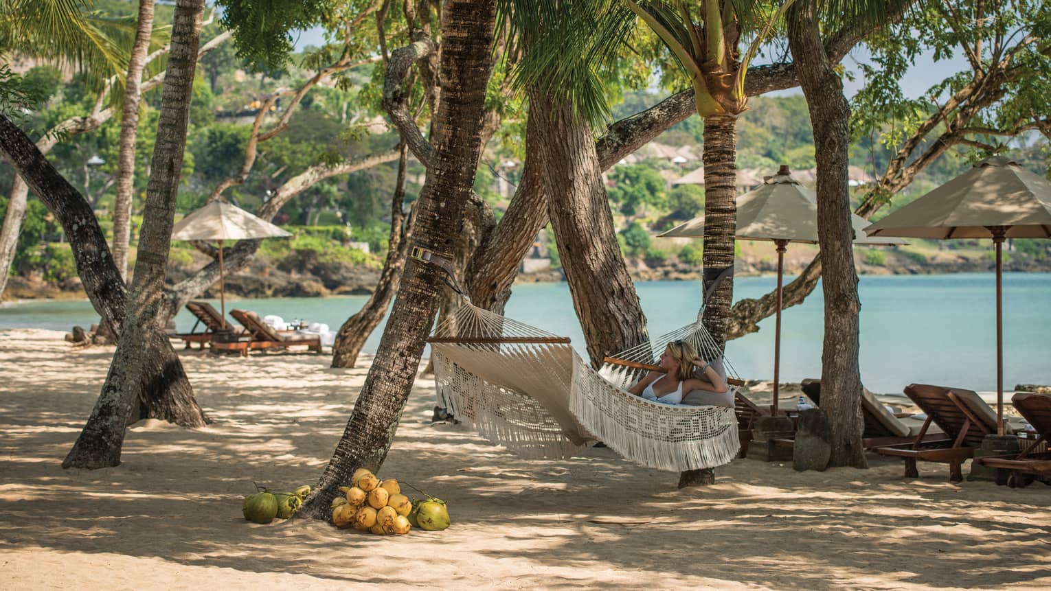 Woman in white bikini lies on white hammock in shade of large trees on Coconut Grove Beach