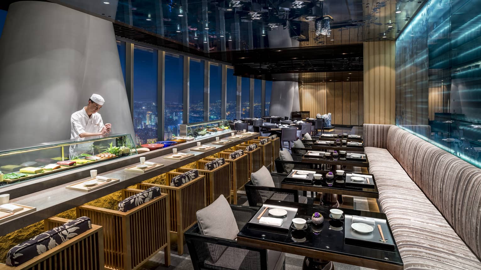 Kumoi Japanese restaurant chef stands behind long sushi bar in dining room overlooking city lights