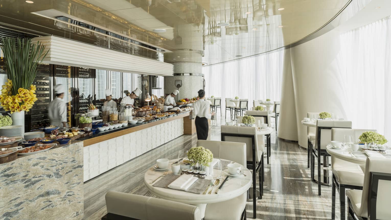 Restaurant dining room with an open live-action kitchen, white walls and chairs, natural light