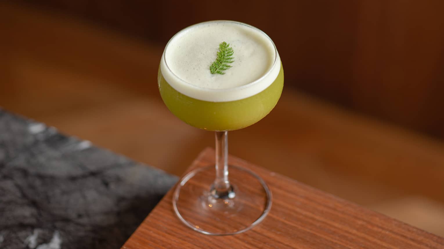 Chartreuese-colored cocktail topped with cream-colored foam and a spring of greenery served in a coupe glass