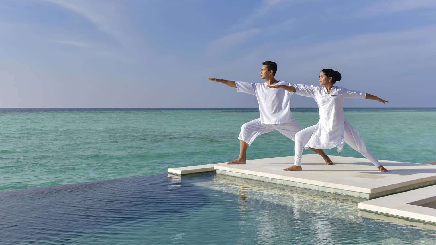 A couple dressed in all white does yoga on a white dock over the sea
