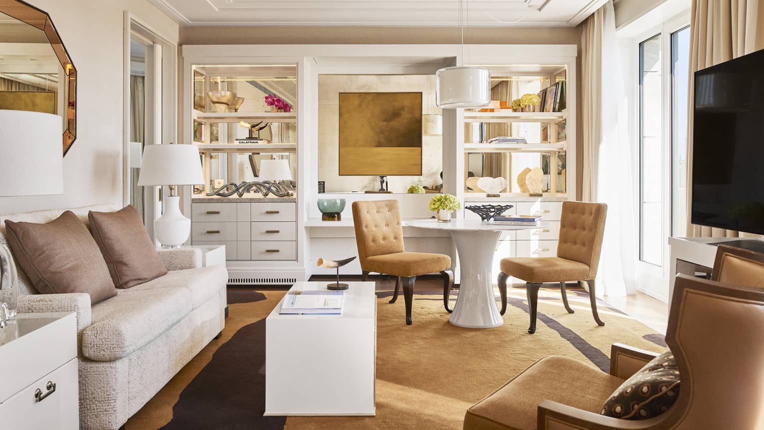 Luxury hotel living room with plush seating, tan chairs, round and rectangular tables, TV, decorative shelves and large windows with beige curtains