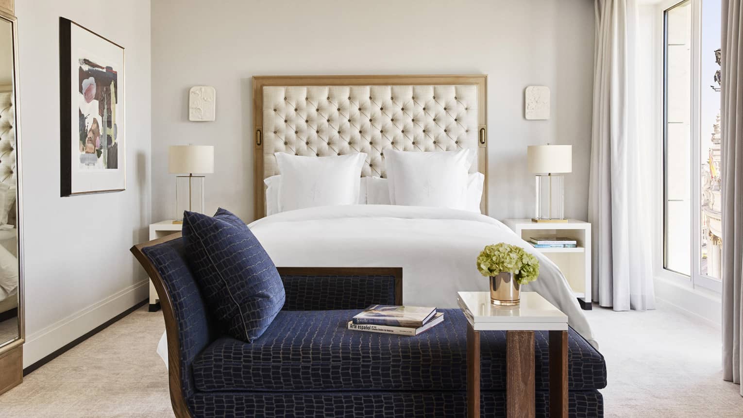 Luxury hotel bedroom with king bed, tufted headboard, blue chaise lounge with books, bedside tables with lamps, wall art and large windows