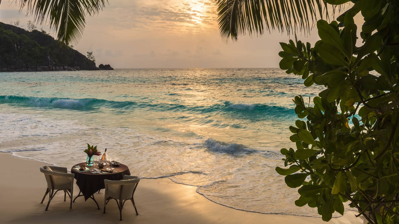 Table for two on sandy beach by ocean's edge at sunset framed by palm trees