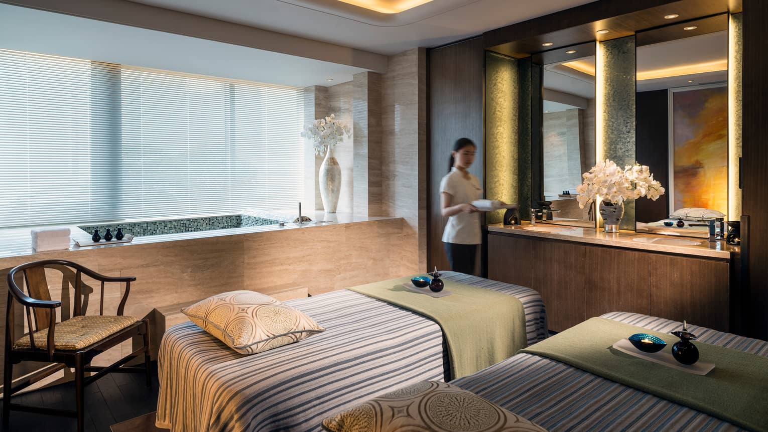 Spa staff carries tray into Couples Massage room with beds, tub