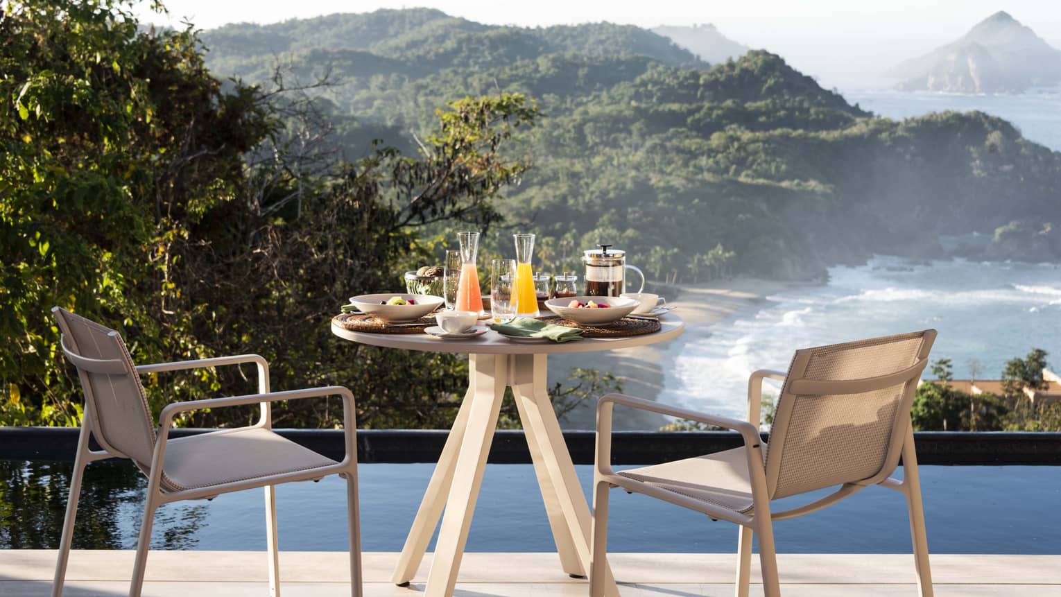 A small table with drinks and food on it looking out a beach and trees.