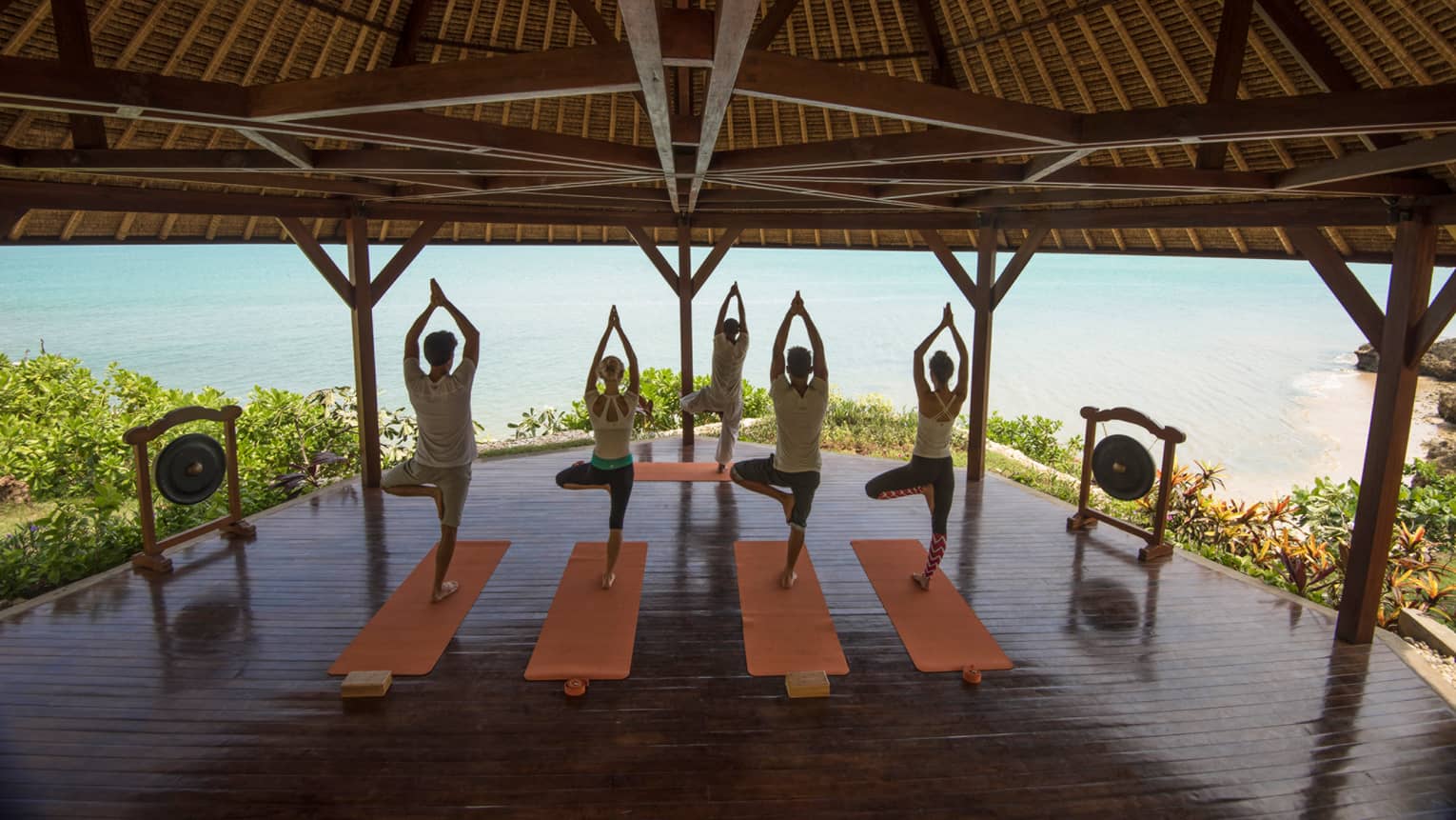 Four people stand in yoga poses, on mats in front of instructor under wood gazebo near ocean