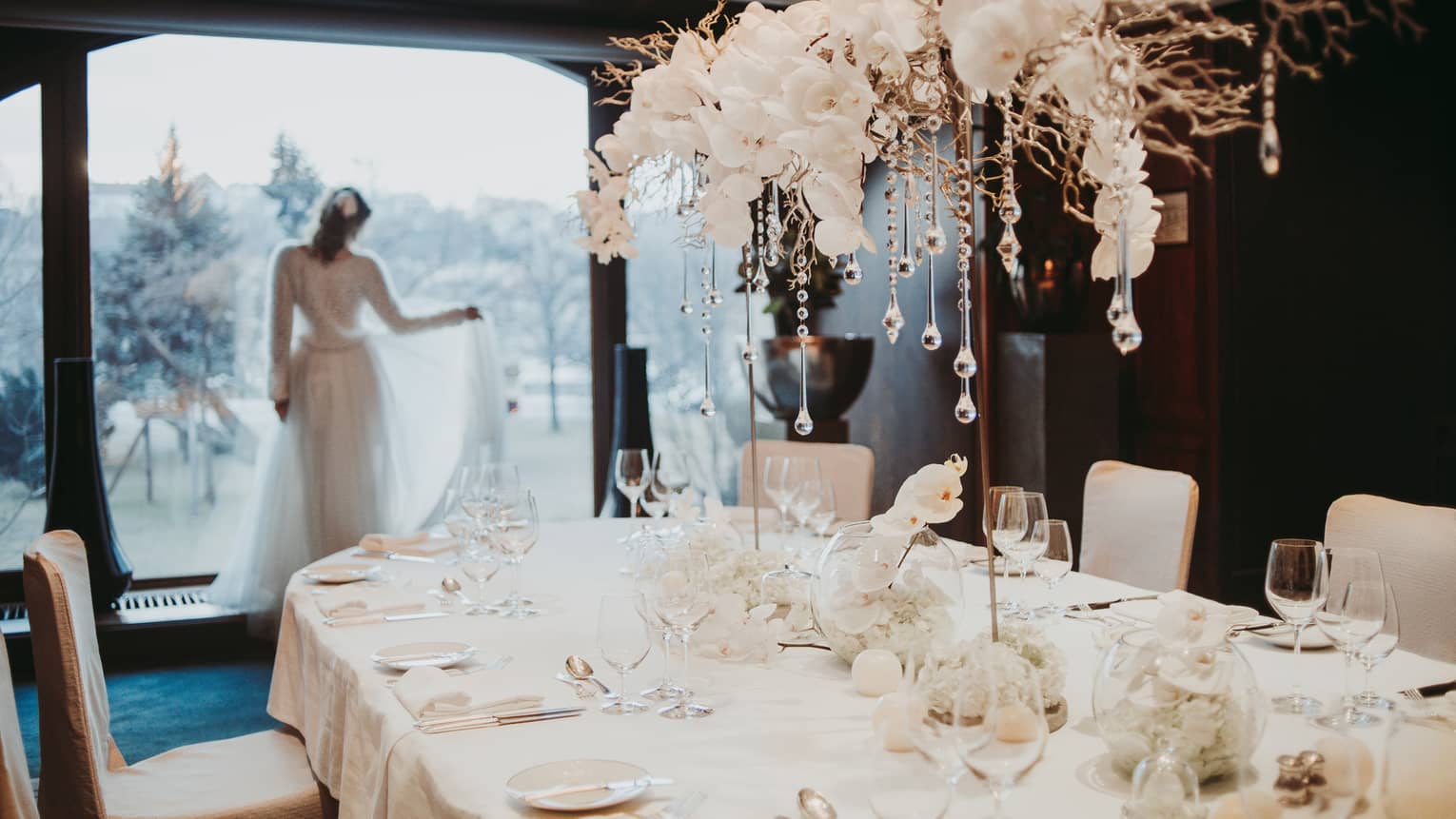 Bride holds train staring out window behind table with white orchids and glass icicles