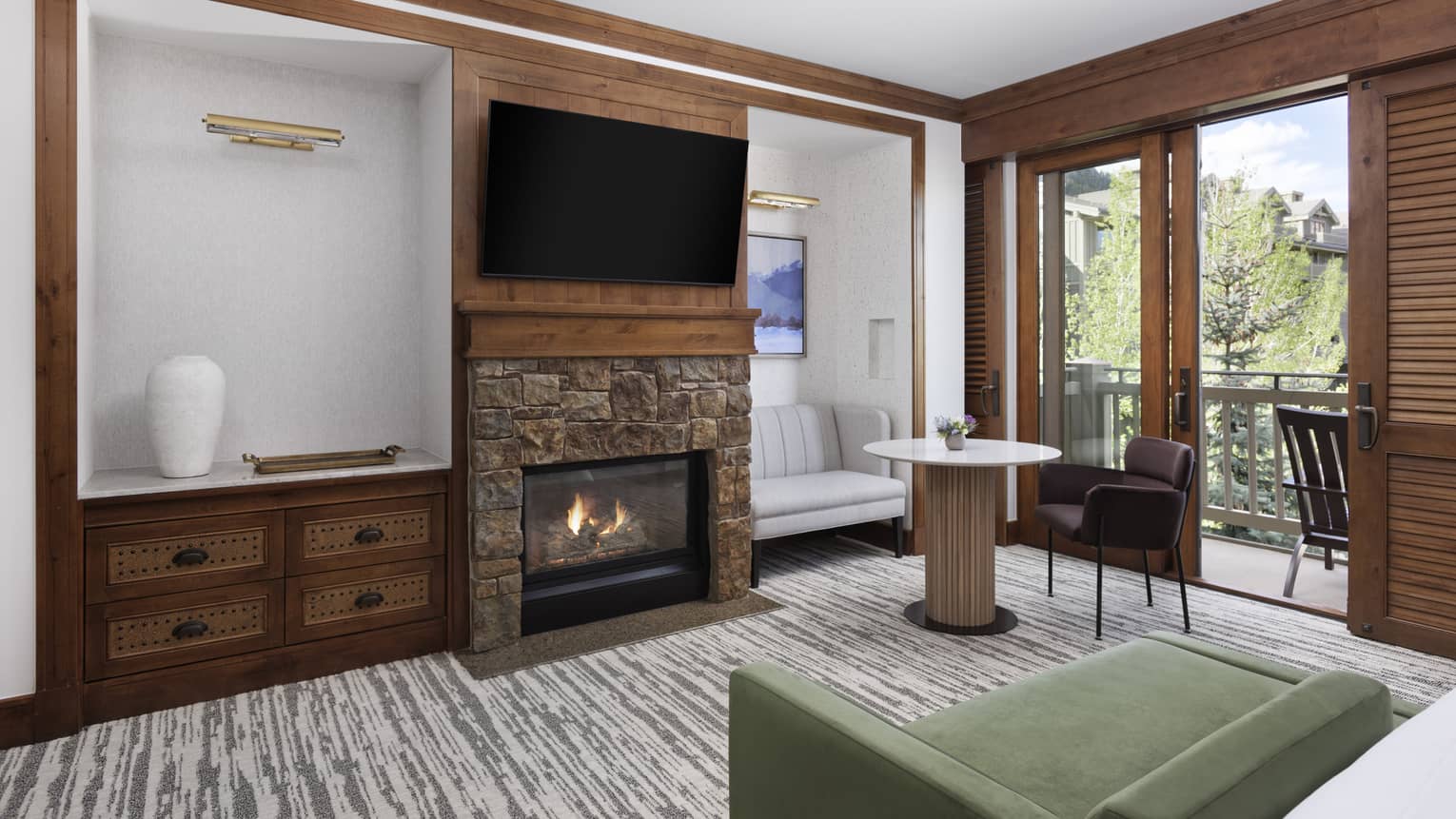 Luxury hotel room lounge area with a stone fireplace, mounted TV, green chaise, sofa and chair by the table, and a balcony with tree views
