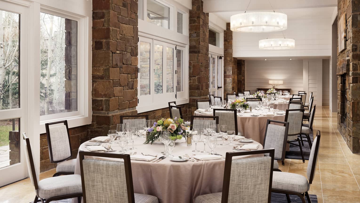 A room with round tables next to large windows, flowers and table settings and grey chairs.