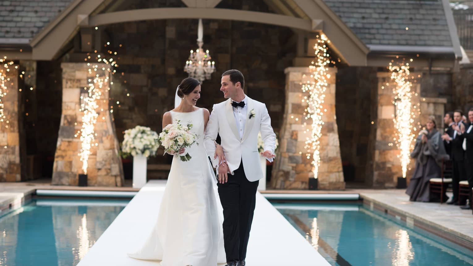 Bride and groom walk along runway across swimming pool as wedding guests clap, fireworks