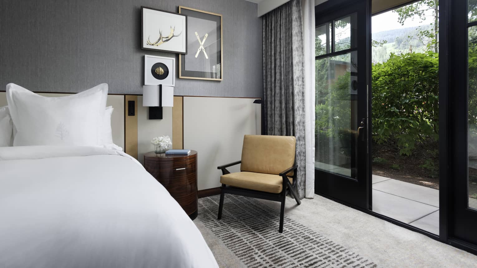 Guest room with bed, white linens, grey walls, wooden side table, lounge chair, sliding glass doors leading to terrace