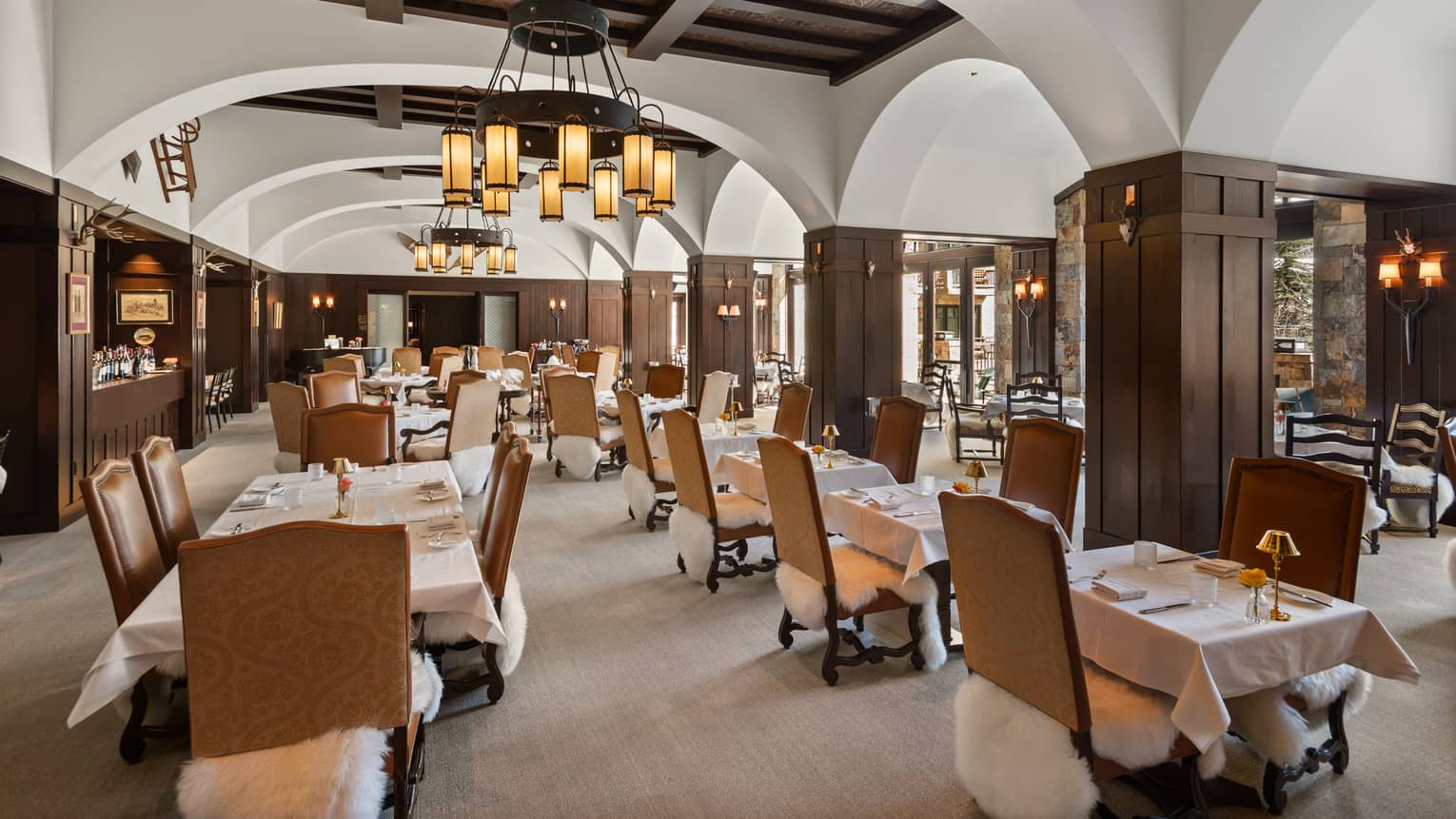 The interior of a well lit restaurant with wood paneling, large white cloth tables and chairs.