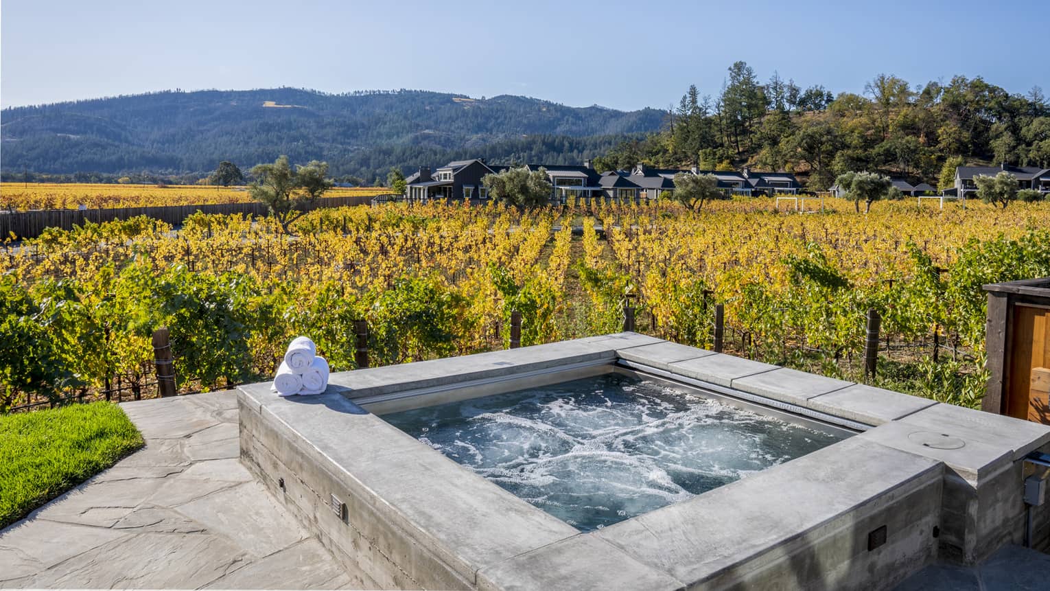 Outdoor hot tub overlooking vineyard rows, surrounded by mountains and luxury resort villas under a clear blue sky