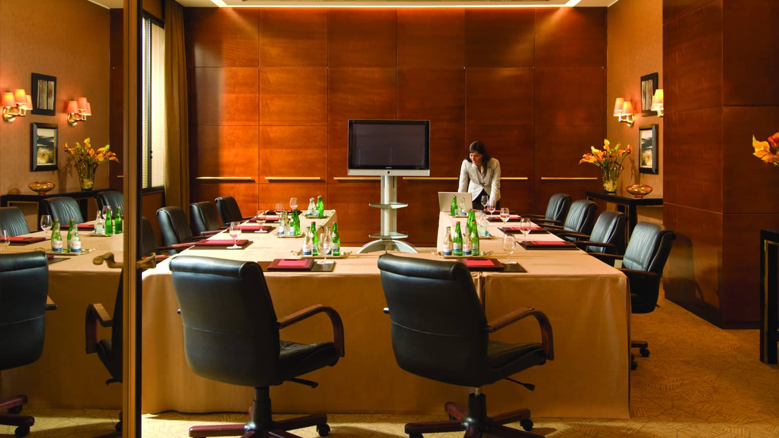 Woman stands in front of U-shaped meeting table set with folders, bottles of water, lined with black office chairs