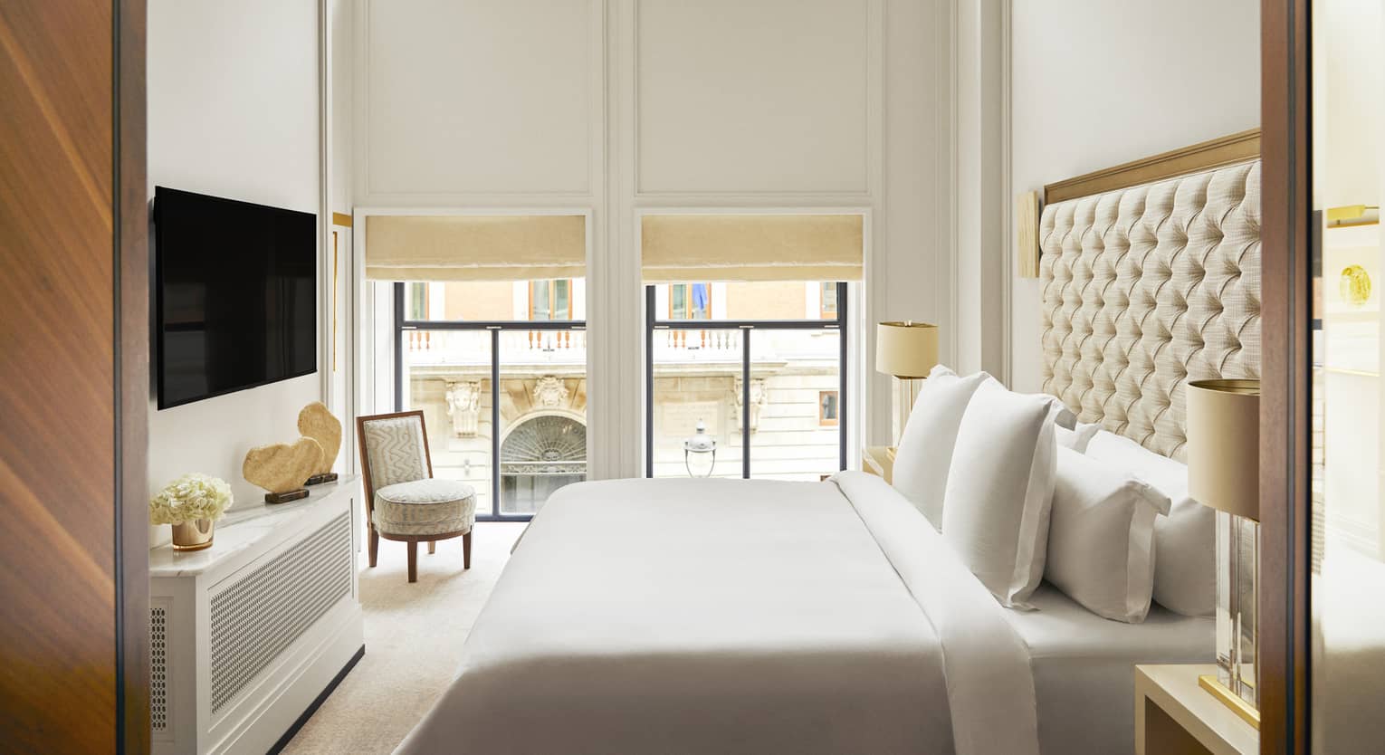 Hotel bedroom with a king bed with tufted headboard, wall-mounted TV, decorative console with stone sculptures, armchair and expansive windows with street views