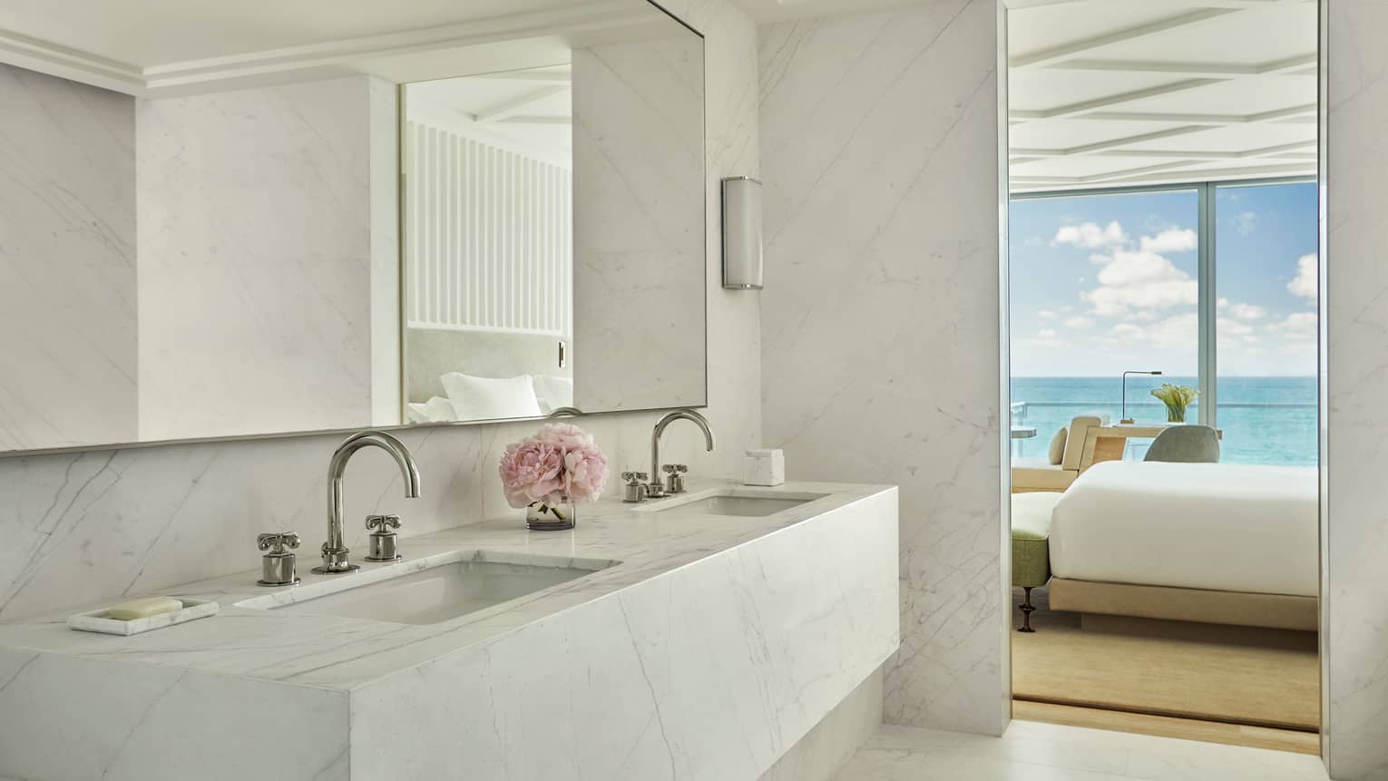 Luxury hotel bathroom with a marble double sink vanity, a mirror and a view into the bedroom with ocean views through floor-to-ceiling windows