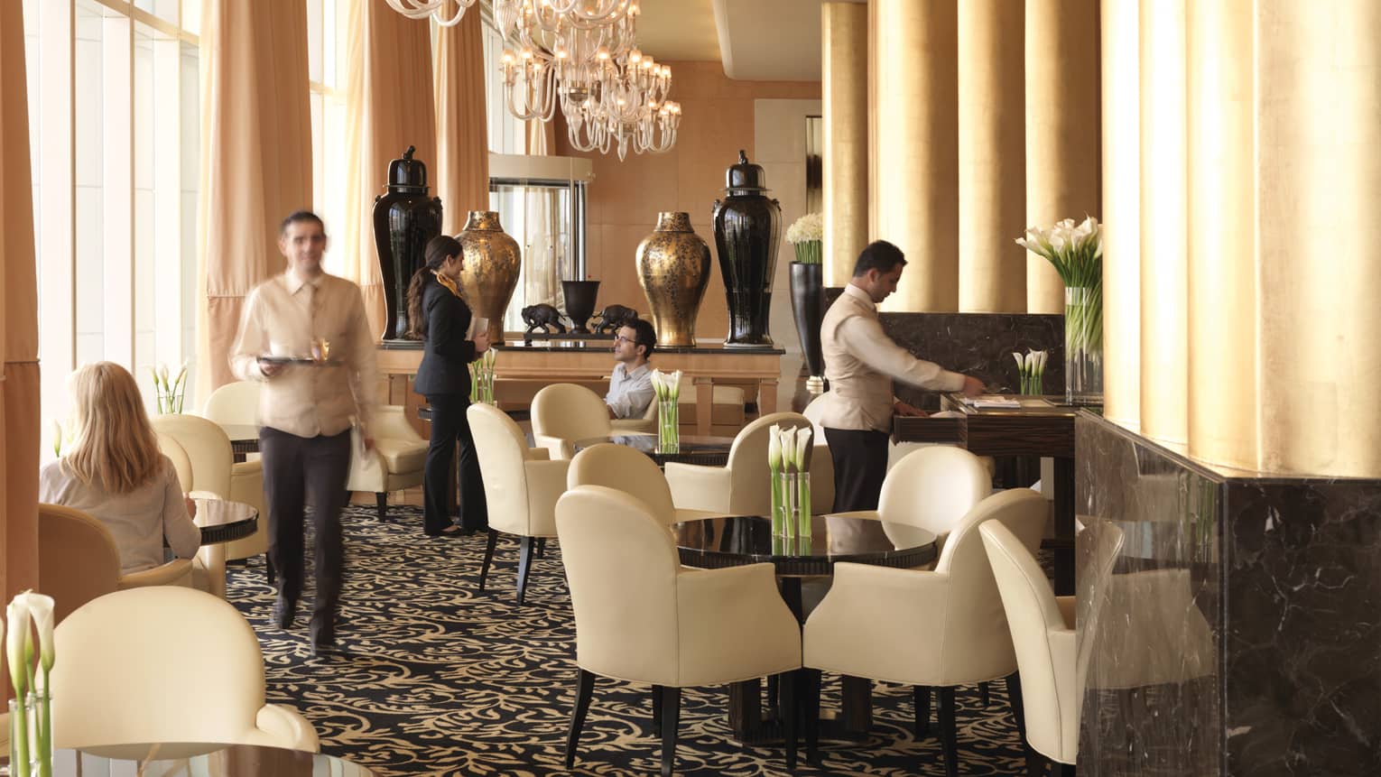 Sunny lobby lounge, hotel staff talk to guests at tables with white leather dining chairs, big gold and black vases behind