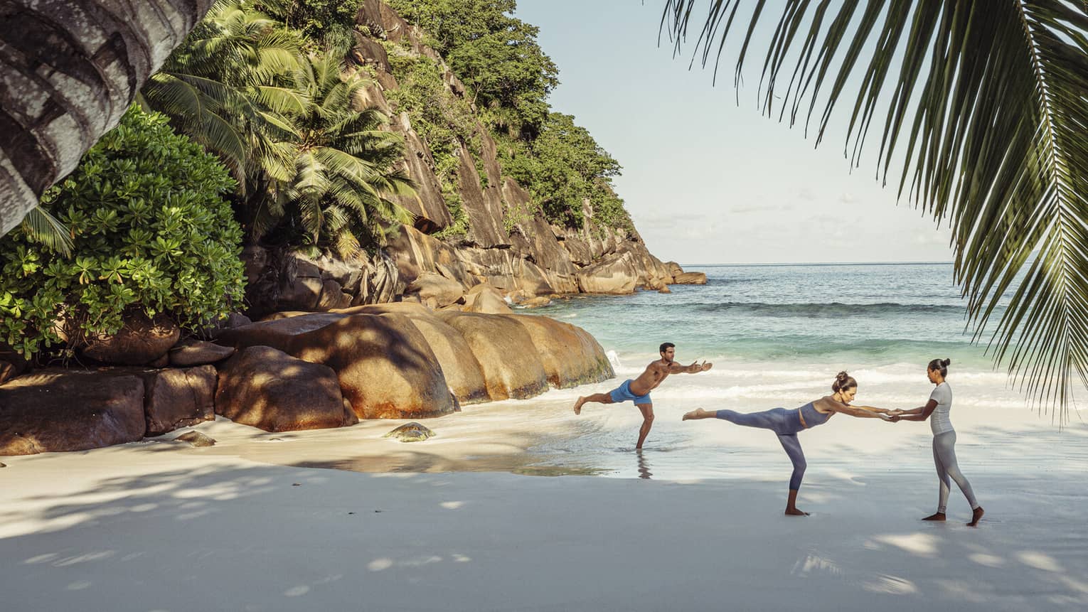 Two guests and an instructor doing yoga on a beach shaded by a large palm tree