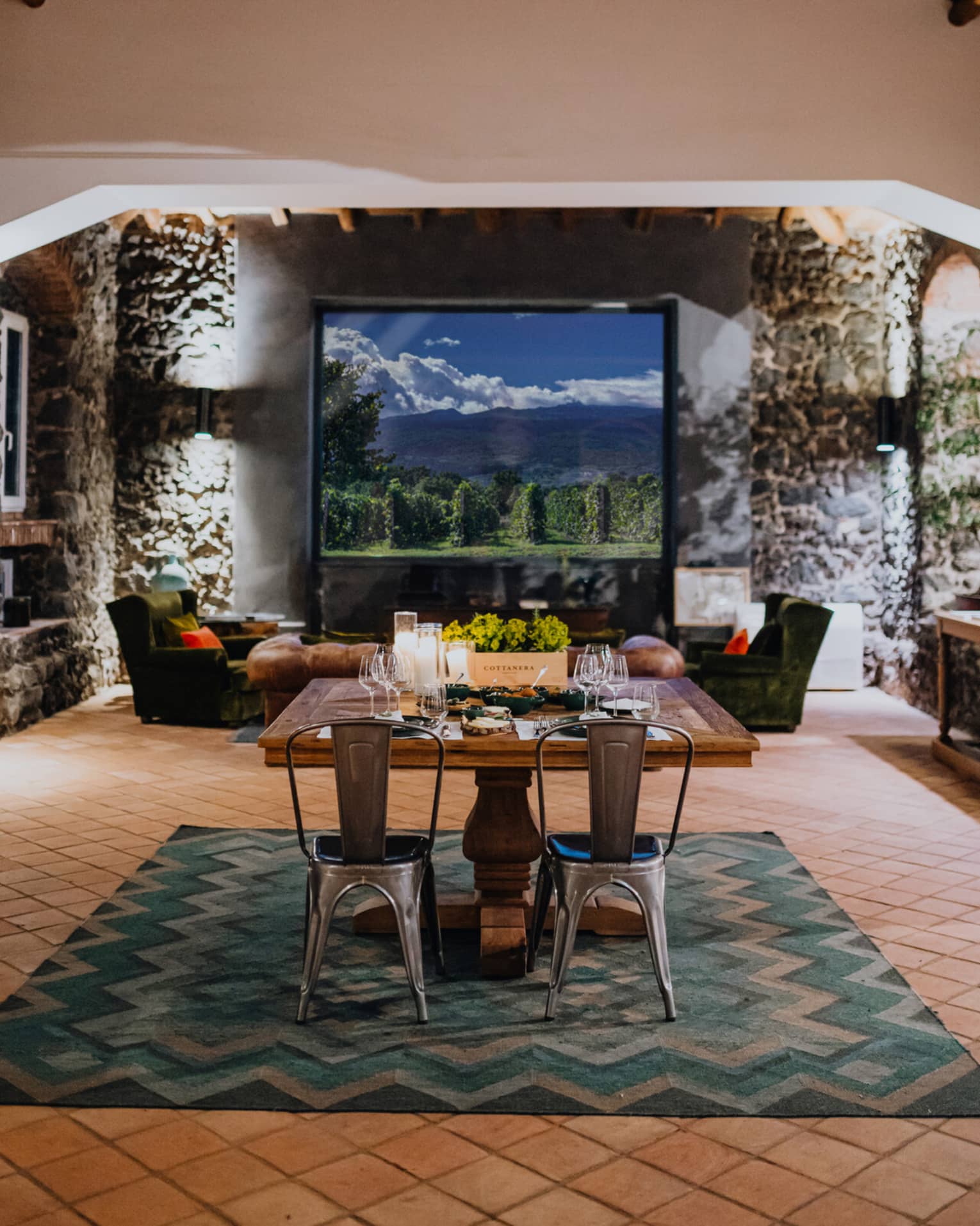 A wooden dining table set with floral arrangements and glassware, in a room with a window overlooking mountains and a vineyard