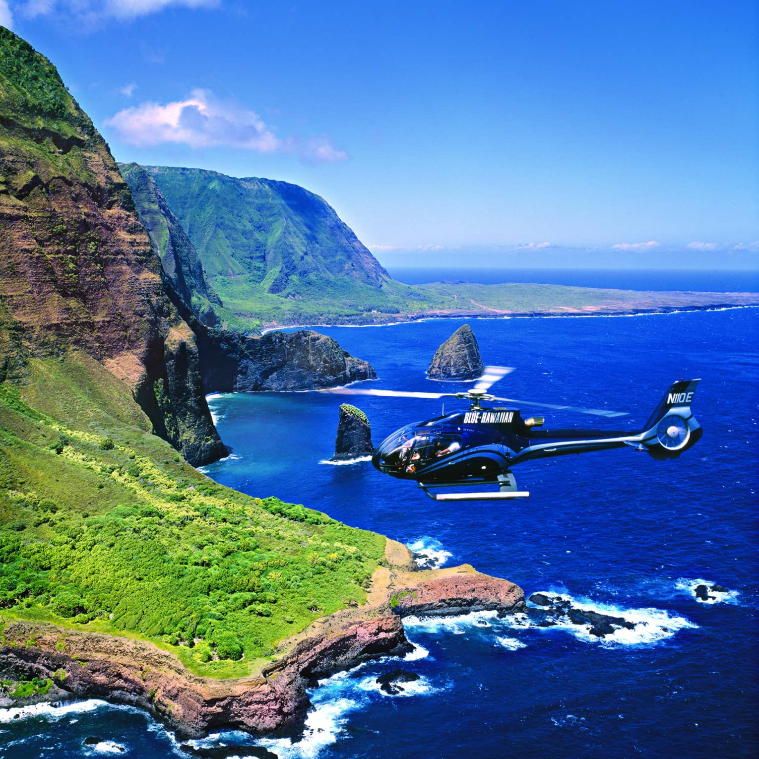 Helicopter flies over ocean and sweeping green cliffs