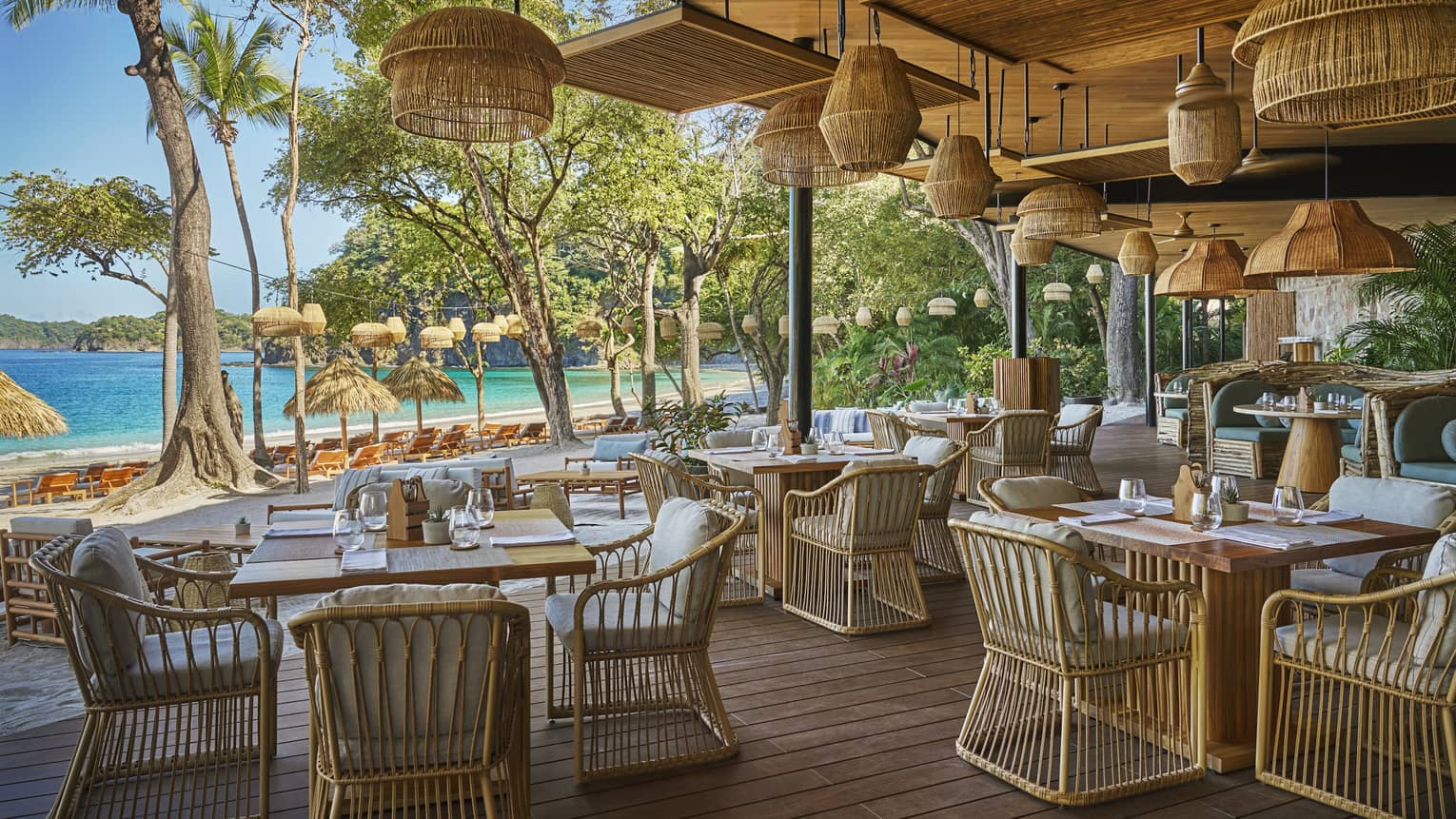 Interior of restaurant dining terrace with multiple dining tables, ratan chairs, rattan light fixtures and a clear view of the nearby beach