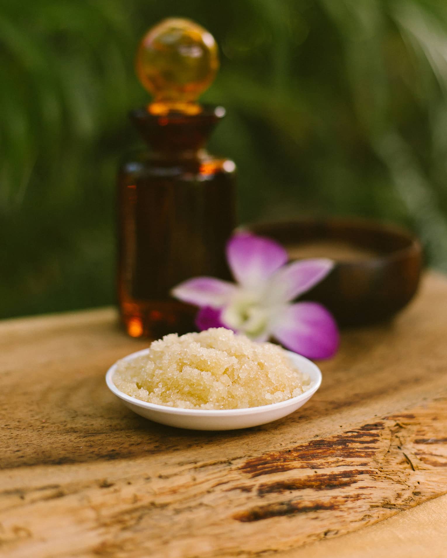 Bowl with salt scrub next to purple tropical flower and glass oil bottle on rustic table
