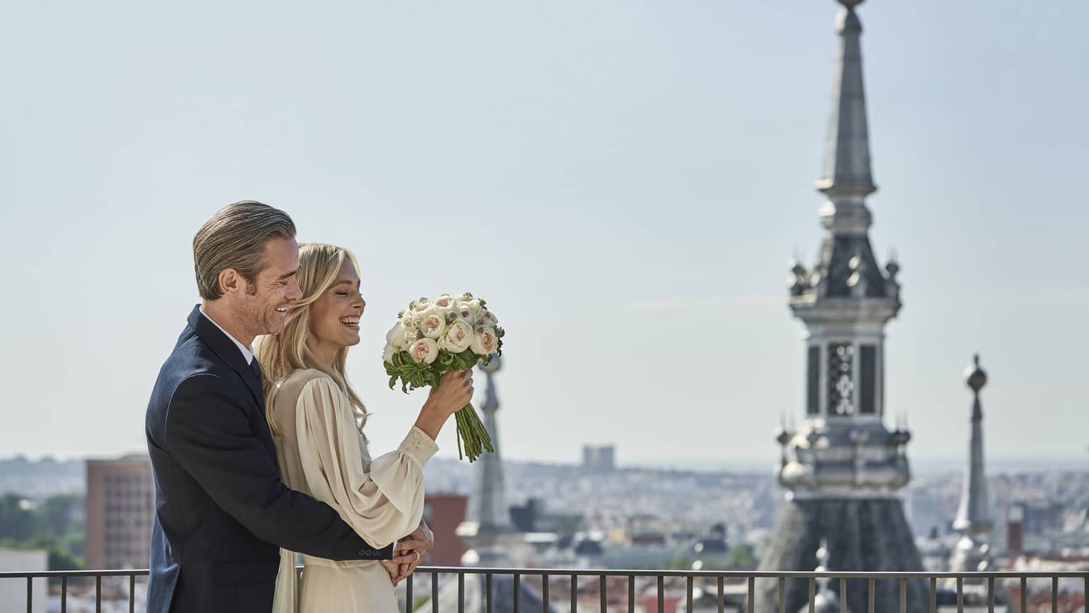 A joyful wedding couple embraces on a rooftop, with the woman holding a bouquet of flowers. The city skyline and a church spire are visible in the background.