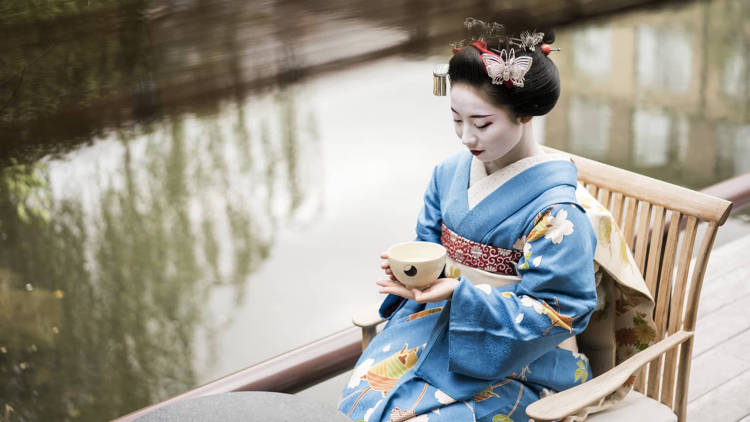Geisha wearing traditional kimono robe, makeup sits on wood chair, holds tea cup