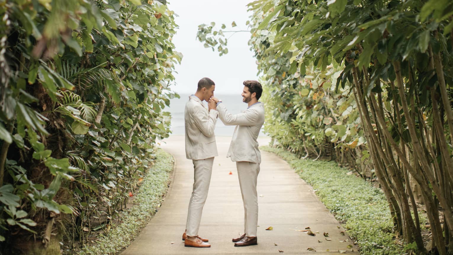 Two men are standing on a pathway of trees and plants while one of the men kisses the others hand.