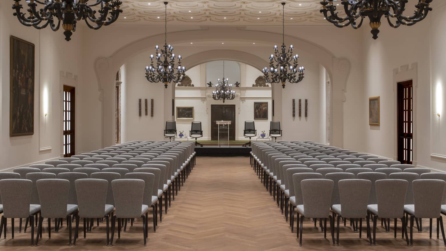 A spacious, neutral-toned space with rows of chairs, a stage area at one end and a textured ceiling, softly lit by elegant chandeliers, recessed ceiling lighting and wall sconces.
