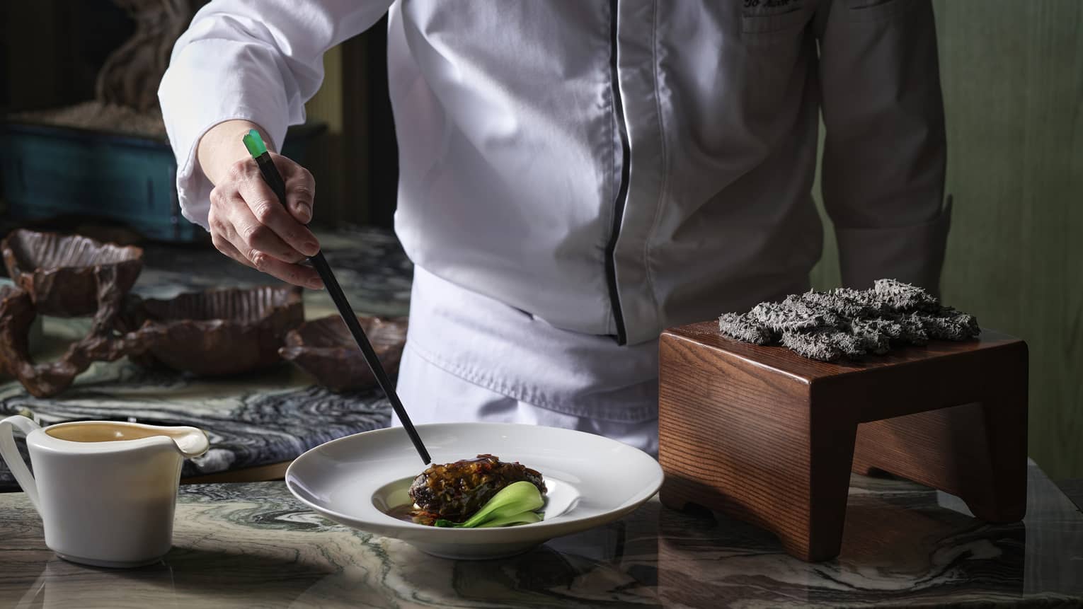 Chef using chopsticks to plate black pork belly in a white dish near a dozen black spiky delicacies on a small wood platform.