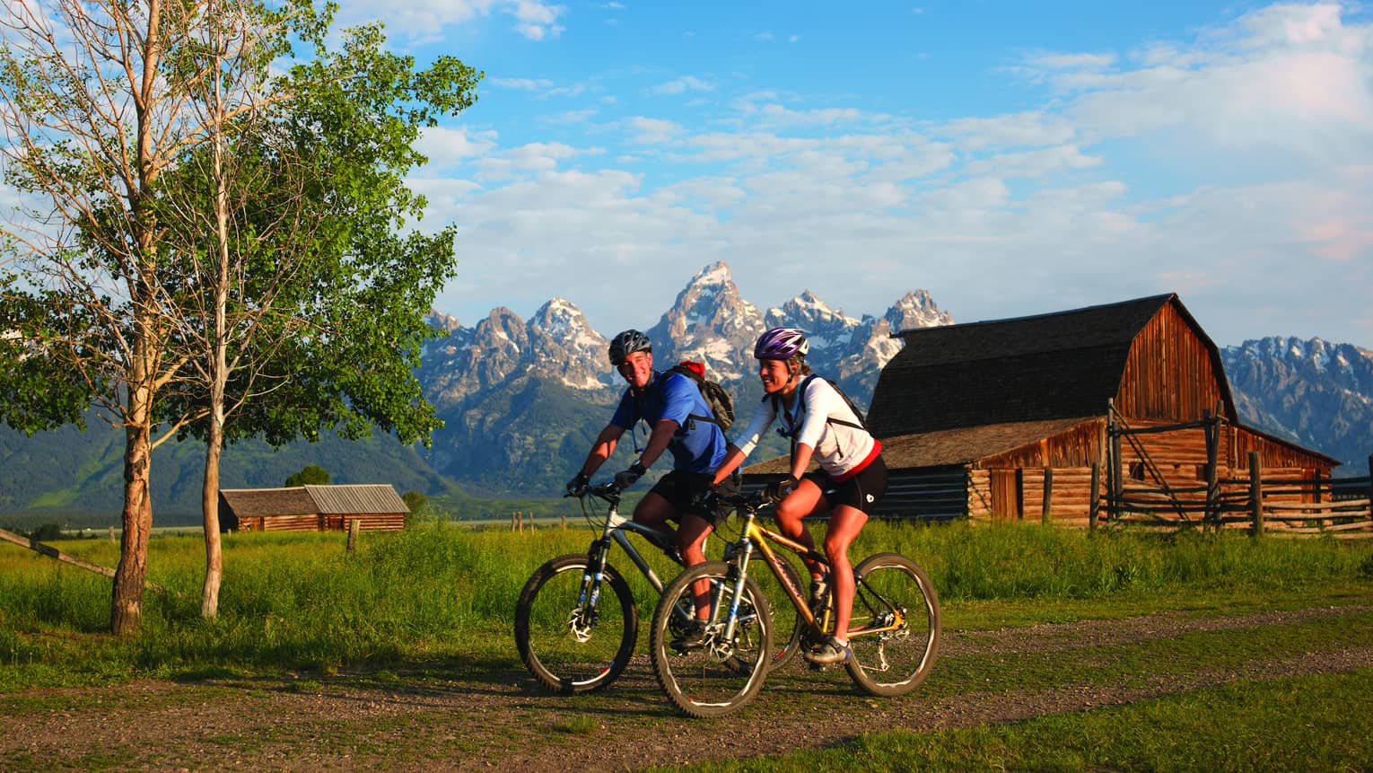 Man and woman on mountain bikes ride on path with log cabin and snow capped mountains in background
