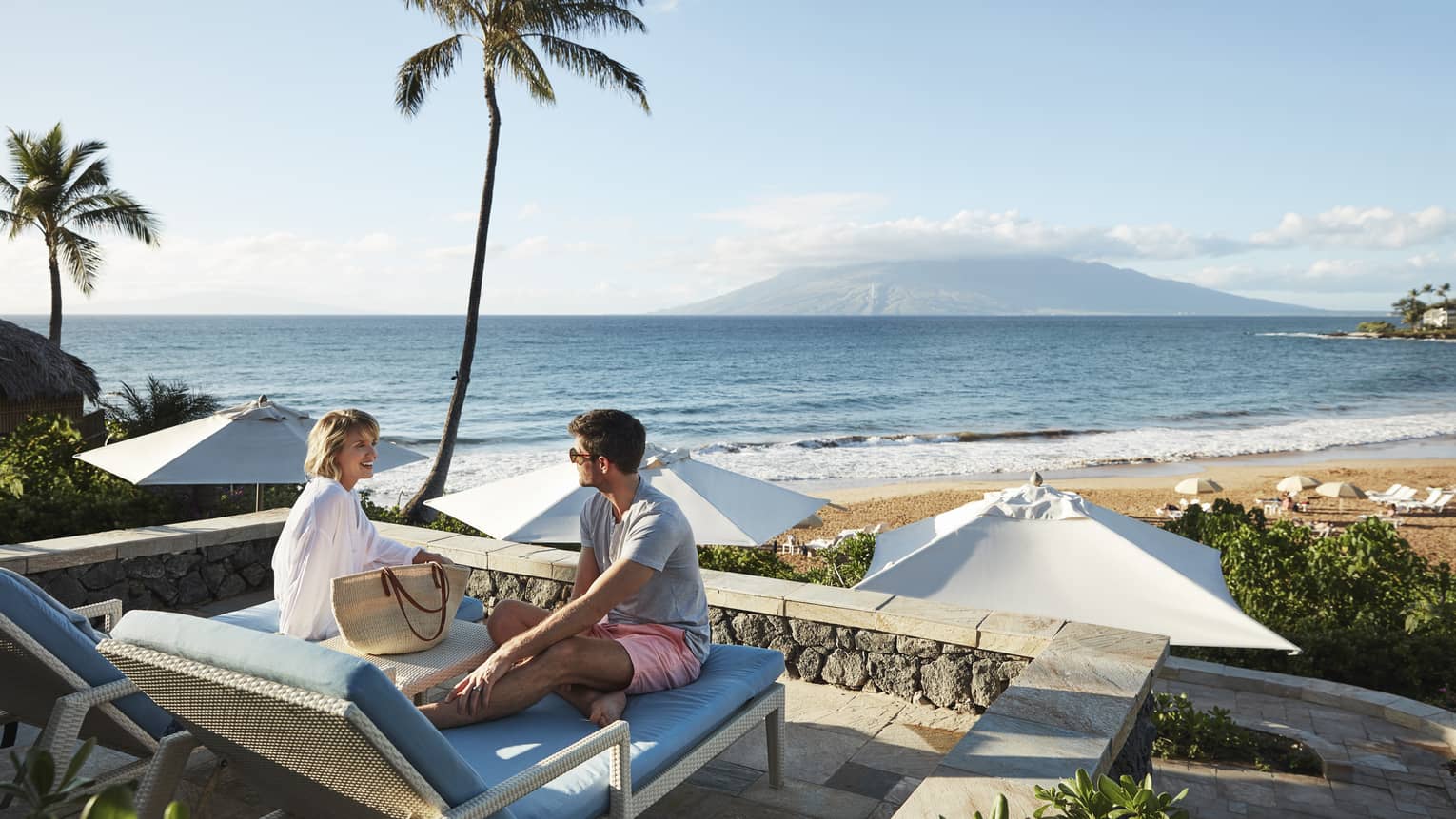 Man in shorts and smiling woman in beach shirt sit on edge of lounge chair, beach and ocean in background