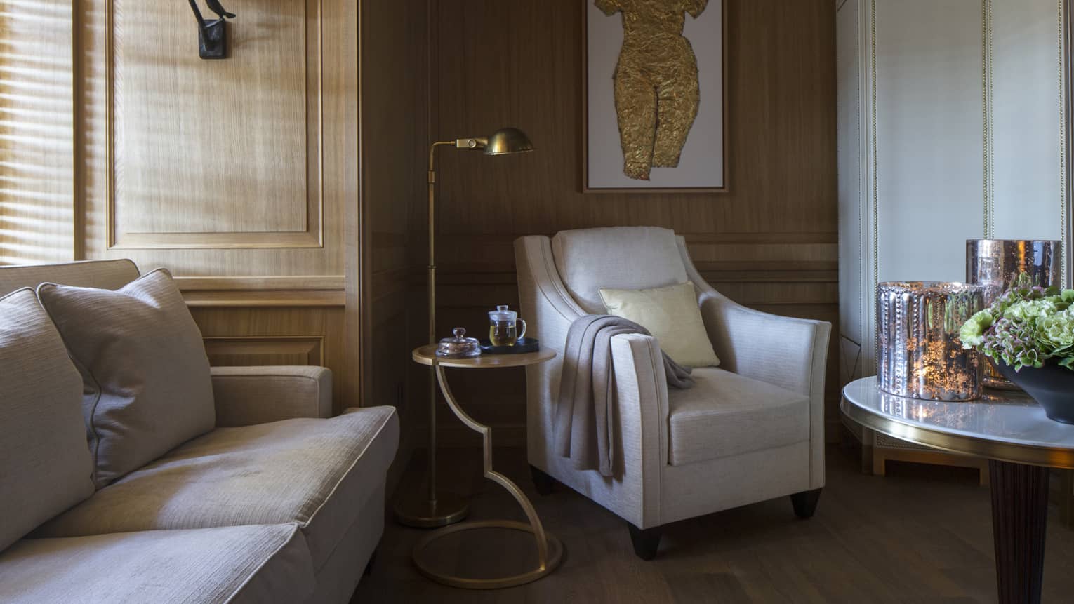 Sunlight streams through window to corner of sofa, white armchair with blanket on arm, reading lamp, art piece on wall