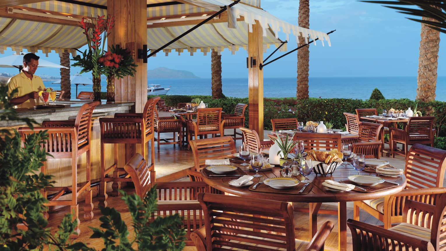 Bartender with colourful cocktails behind large round wood patio tables at Reef Grill at dusk