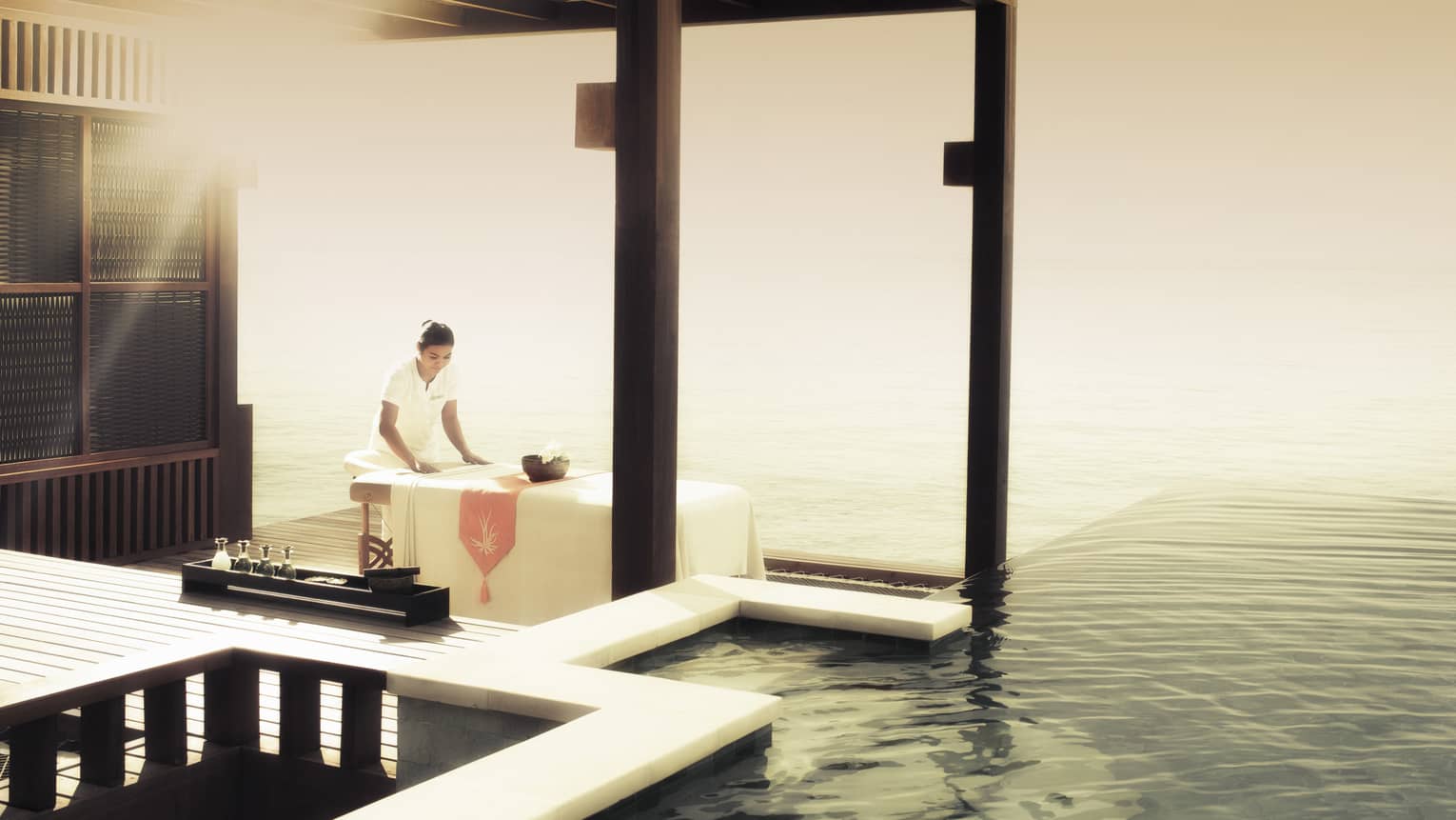 Spa therapist preparing a massage table in a serene overwater pavilion with an ocean view at sunrise