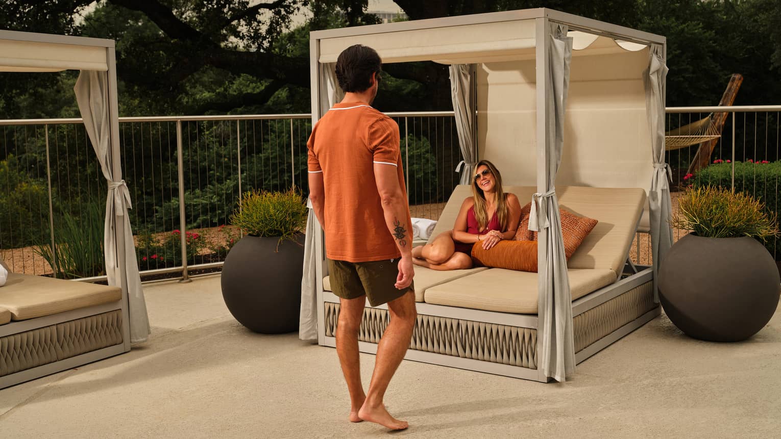 A man walks towards a woman lounging in a cabana surrounded by large potted plants