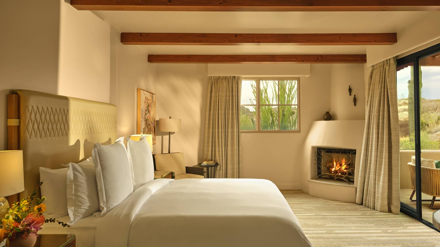 Light streams into a bedroom of luxury suite, with a king bed, fireplace, beamed ceiling and walk-out terrace