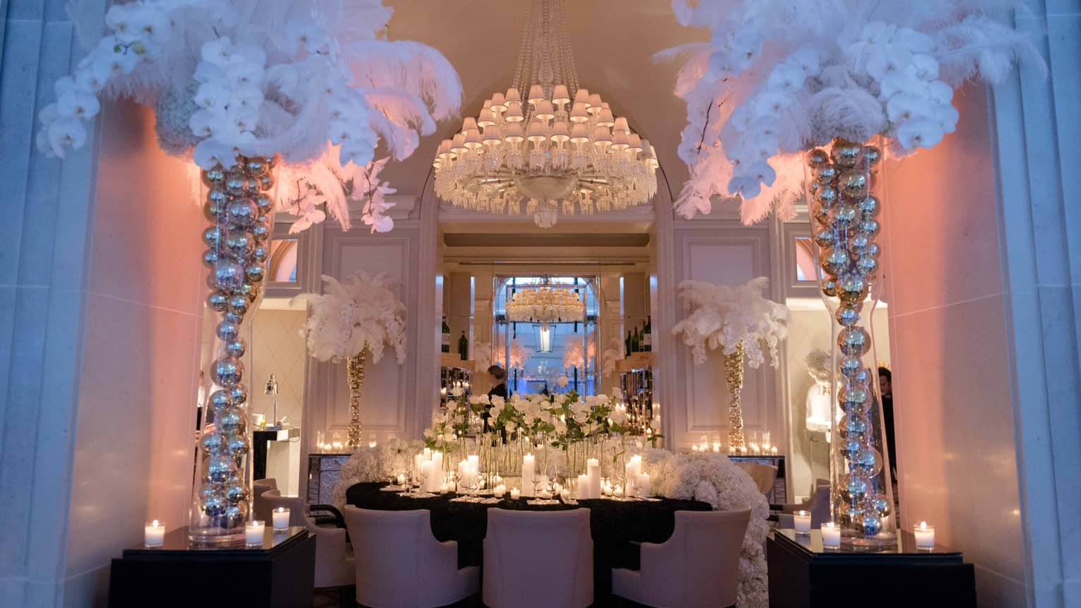 Dining table under crystal chandelier, surrounded by tall vases filled with silver balls, faux white palms