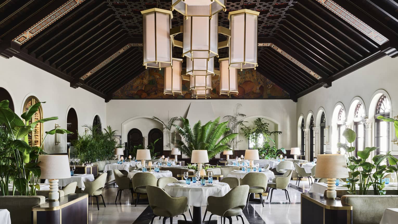 A sophisticated dining room with high, dark wood-beamed ceilings, large hanging lantern chandeliers, green upholstered chairs, white tablecloths and indoor plants.