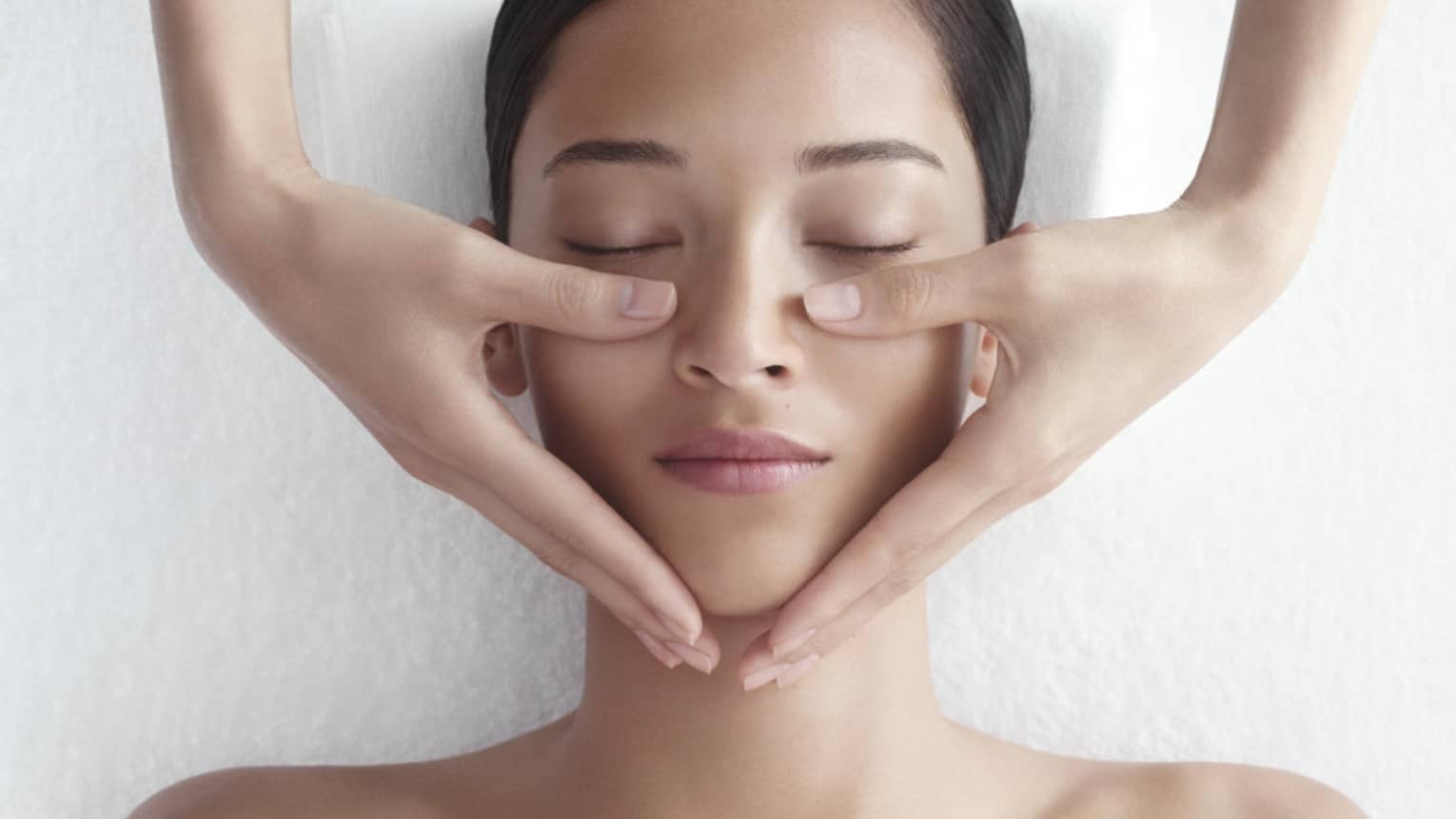 Woman in white bathrobe lies on massage table with eyes closed, masseuse rests hands on her cheeks