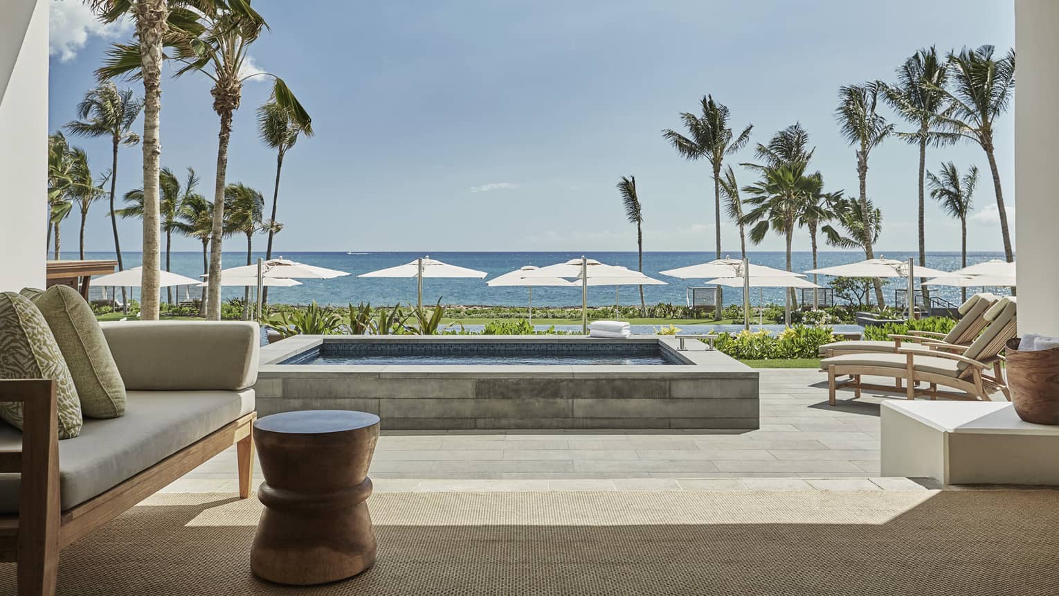 Pacific Suite's luxurious beachfront patio with a comfortable seating area, a private pool and sun loungers facing the ocean, framed by palm trees and umbrellas under a clear sky