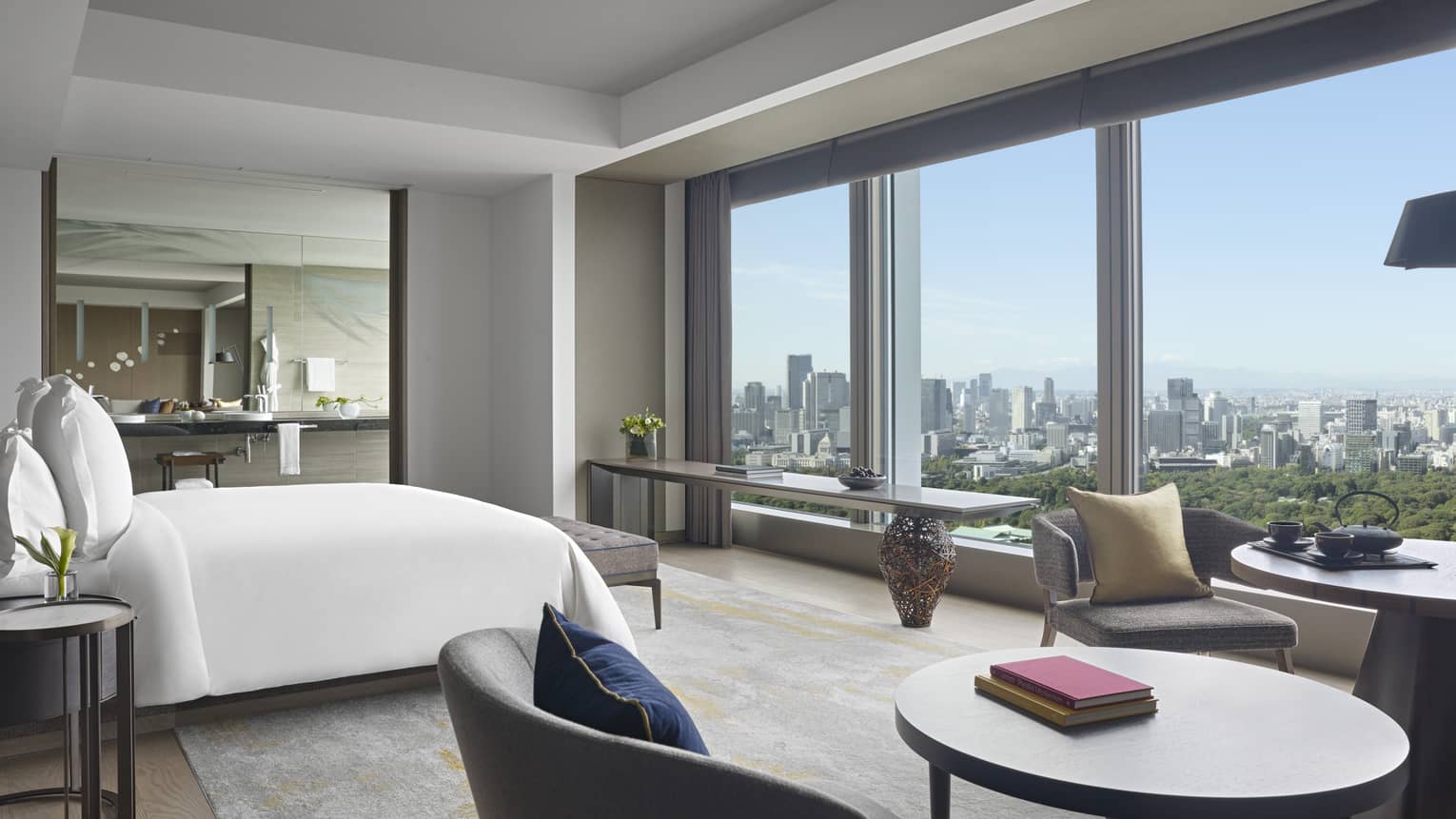 Hotel room with chair, table and king bed facing wall of windows overlooking Tokyo gardens and city