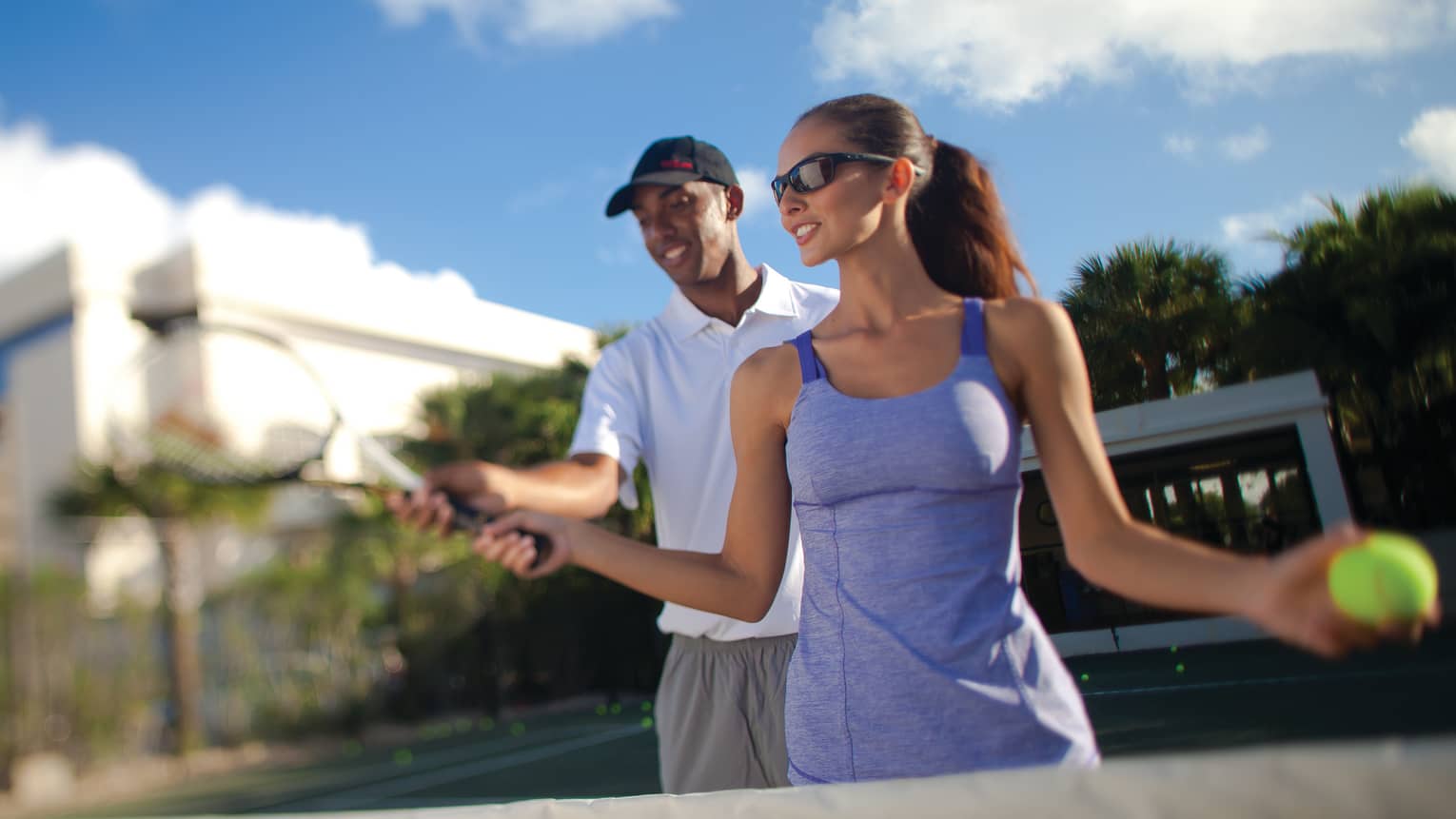 Man helps woman with tennis racket on court under blue sky