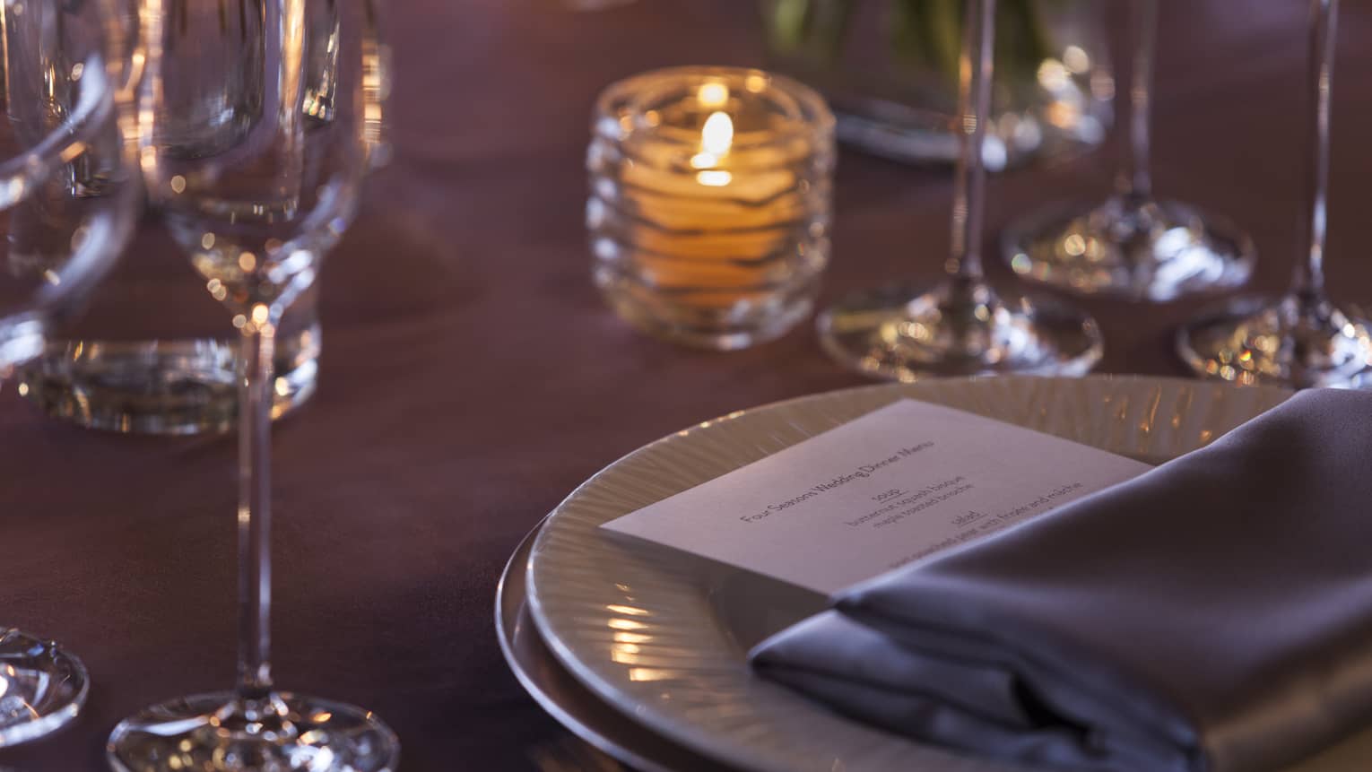 Close up of table with candle, grey napkin and dinner menu on white plate, glasses