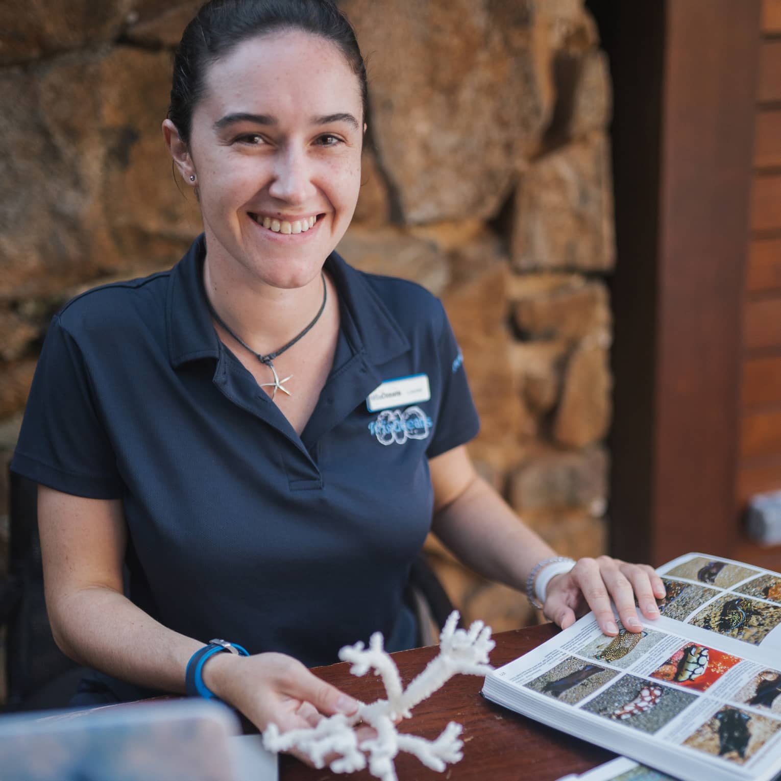 A smiling marine biologist reads an illustrated book of sea creatures, a piece of white coral held in their other hand.